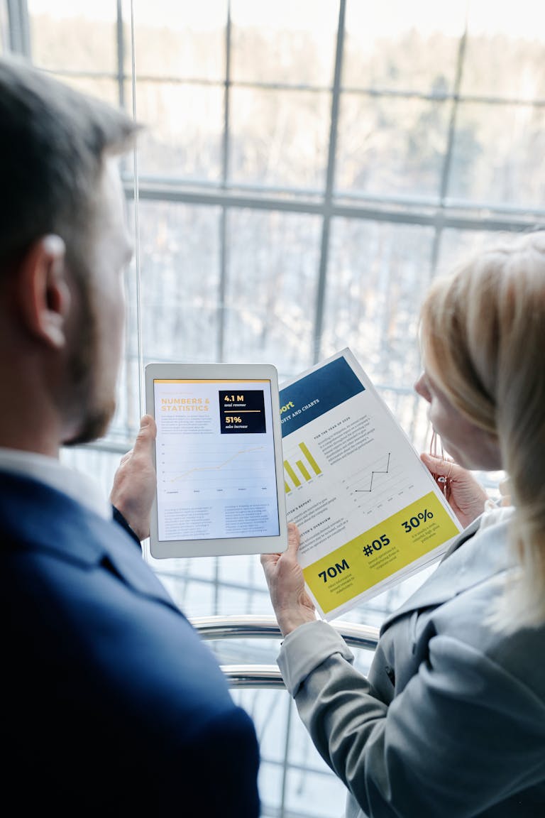 Two business professionals reviewing graphs and data on a tablet and paper report indoors.